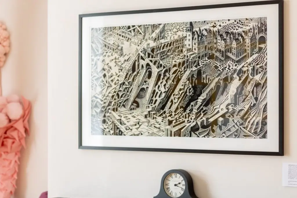 Entropy Drift, a monochrome print of a crumbling, Escher-like cityscape, framed on a wall above a mantel clock.