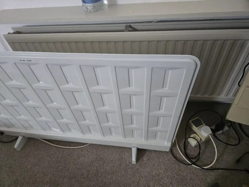 A portable electric panel heater standing in front of a radiator, plugged into an energy meter.