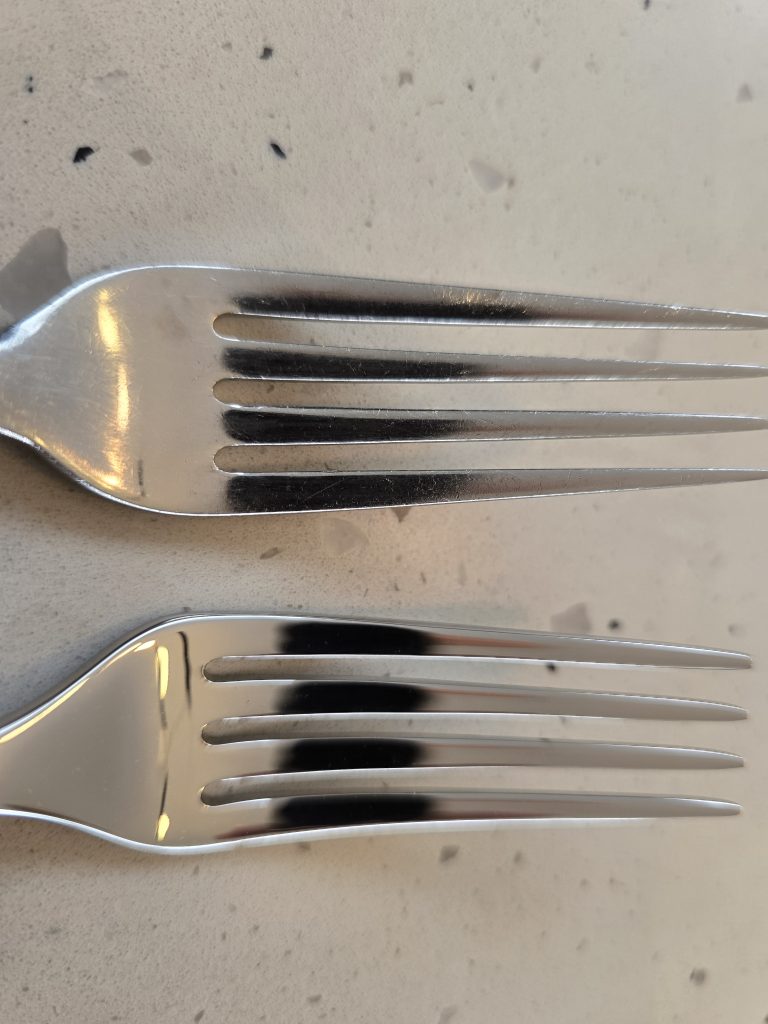 An older Robert Welch Radford set of fork tines beside a new one, on a speckled worktop.