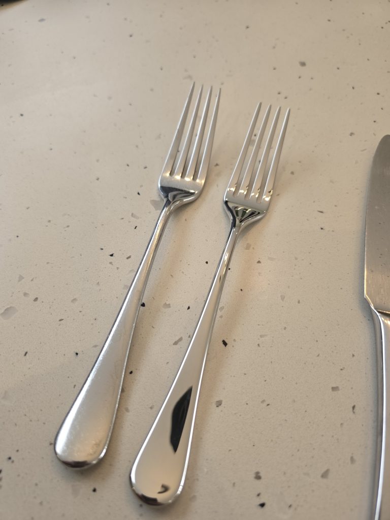 An older Robert Welch Radford table fork beside a new one, on a speckled worktop.