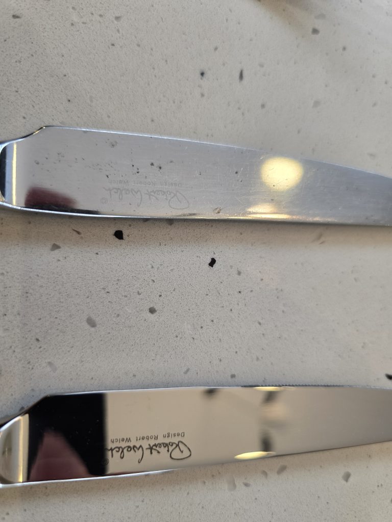 An older Robert Welch Radford knife blade, showing the engraved logo, beside a new one, on a speckled worktop.