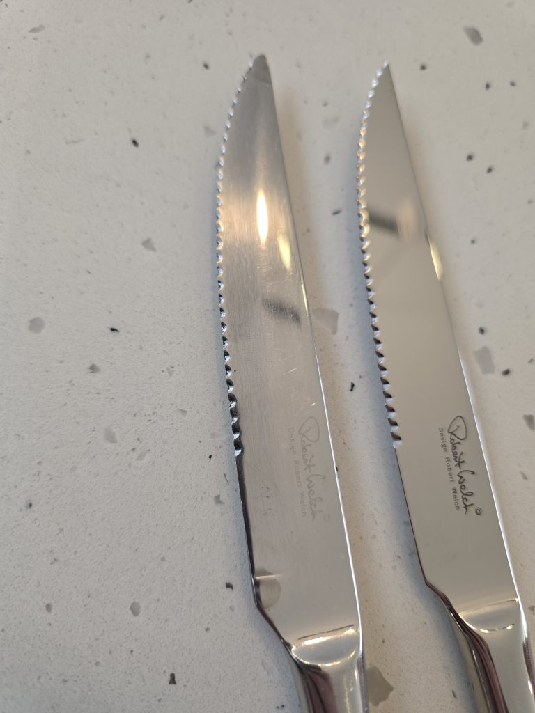 An older Robert Welch Radford serrated steak knife beside a new one, on a speckled worktop.