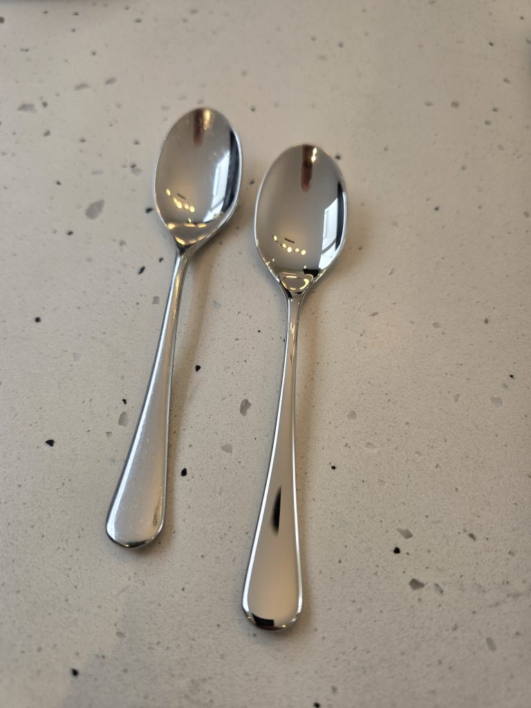 An older Robert Welch Radford teaspoon beside a new one, on a speckled worktop.